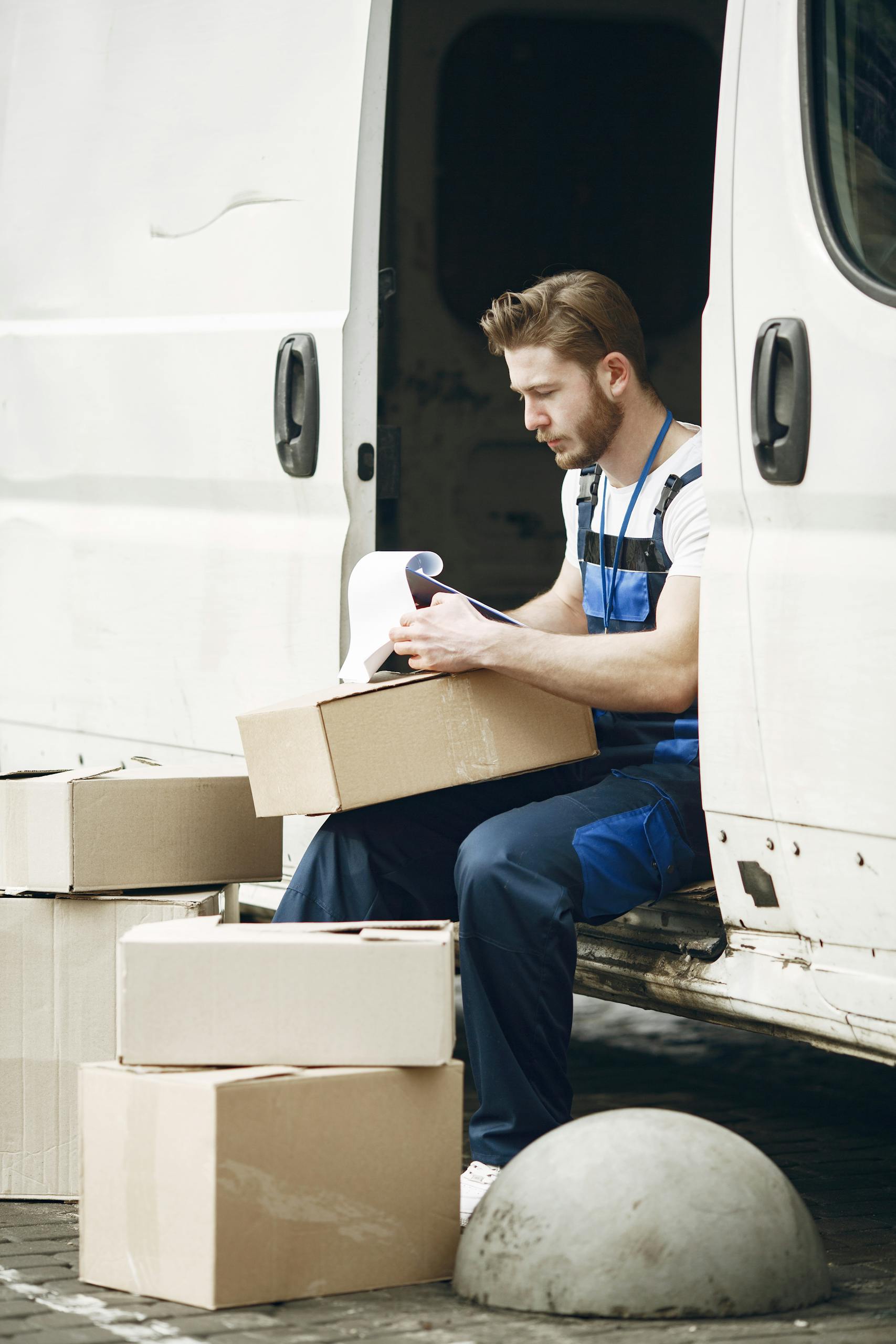 Deliveryman in overalls sorting packages next to a white van outdoors.